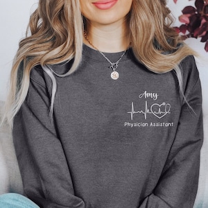 Könnte beinhalten: Ein dunkelgrauer Pullover mit einem weißen Design, das einen Herzschlag, ein Herz und ein Stethoskop zeigt. Der Text "Amy Physician Assistant" ist unter dem Design gedruckt.