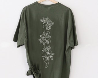 Pode incluir: Uma camiseta verde escura com um contorno branco de uma coluna vertebral com flores crescendo dela.