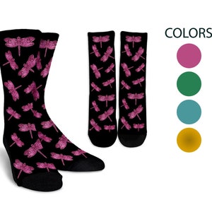 Op de afbeelding: Een paar zwarte sokken met een roze libellenpatroon. De sokken worden getoond tegen een witte achtergrond. Het woord "COLORS" staat in zwarte tekst boven vier gekleurde cirkels: roze, groen, teal en goud.