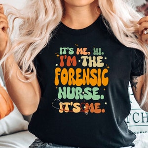 Könnte beinhalten: Ein schwarzes T-Shirt mit einer Grafik im Retro-Stil, auf dem "It's me, hi. I'm the Forensic Nurse. It's me." steht.
