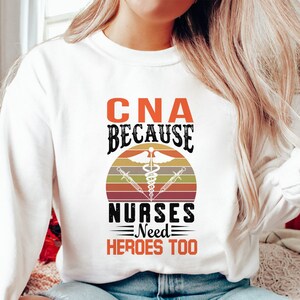 Puede incluir: Sudadera blanca con un gráfico de estilo vintage que dice "CNA because nurses need heroes too". El gráfico presenta un símbolo de caduceo y una puesta de sol.