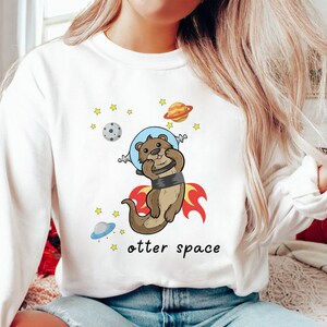 May include: White sweatshirt featuring a cartoon otter wearing a space helmet and flying through space. The text "otter space" is printed below the otter.