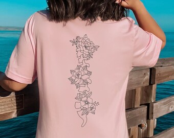 Pode incluir: Uma camiseta rosa com um contorno preto de uma coluna vertebral com flores. A camiseta está sendo usada por uma pessoa em pé em um píer de madeira.