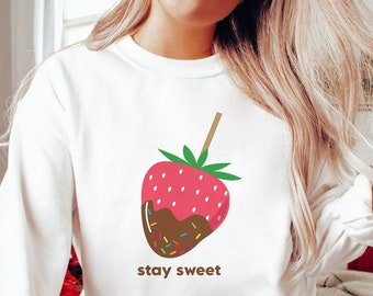 Puede incluir: Sudadera blanca con un gráfico de una fresa cubierta de chocolate en un palo con chispas. El texto "stay sweet" está impreso debajo de la fresa.