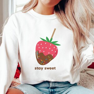 May include: White sweatshirt with a graphic of a chocolate-covered strawberry on a stick with sprinkles. The text "stay sweet" is printed below the strawberry.