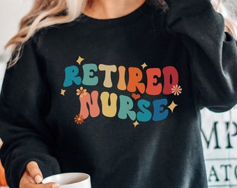 Puede incluir: Una sudadera negra con el texto "Retired Nurse" en letras arcoíris con flores y brillos. La sudadera la lleva puesta una persona sentada en una cama.