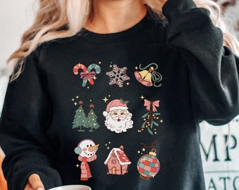 May include: Black sweatshirt featuring a grid of Christmas-themed illustrations. Designs include a candy cane, snowflake, bell, Christmas tree, Santa Claus, snowman, house, and ornament. The sweatshirt is worn by a person holding a white mug.
