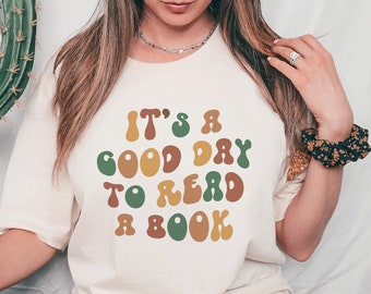 May include: A cream-coloured t-shirt featuring the phrase "It's a good day to read a book" in a retro font. The text is in shades of brown, green, and orange. The t-shirt has a relaxed fit.