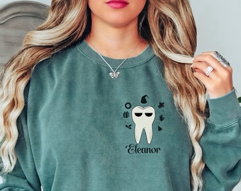 May include: A teal-colored sweatshirt featuring a cartoon tooth wearing sunglasses and a witch hat. The name "Eleanor" is printed below the tooth. The sweatshirt has a crew neck and long sleeves. The words "Blue Spruce" are printed on the bottom right.