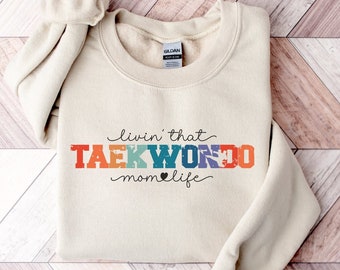 May include: A beige sweatshirt with the text "livin' that TAEKWONDO mom life" in colorful letters with a heart symbol.
