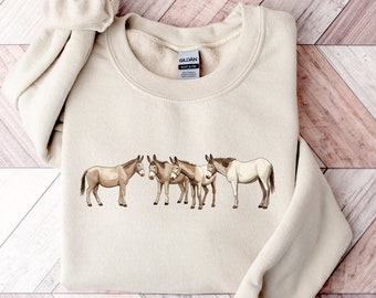 Puede incluir: Una sudadera beige con un gráfico de cinco burros marrones de pie en fila. Los burros están mirando hacia el lado izquierdo de la imagen. La sudadera tiene una etiqueta Gildan en la parte inferior izquierda de la imagen.