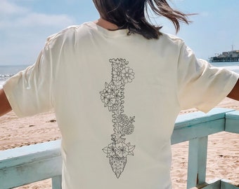 Pode incluir: Uma camiseta branca com um desenho de linha preta de uma espinha dorsal com flores crescendo dela. A camiseta está sendo usada por uma pessoa em pé em uma plataforma de madeira com uma grade azul. O fundo é uma praia com água azul e um píer à distância.