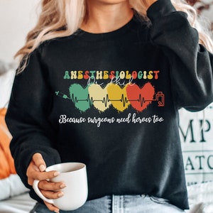 May include: Black sweatshirt with a graphic design that says "Anesthesiologist - Because surgeons need heroes too". The graphic features a stylised heart with a heartbeat line and a syringe.