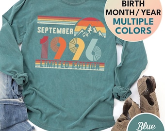 May include: A blue spruce long-sleeve shirt featuring a retro-style graphic with the text SEPTEMBER 1996 LIMITED EDITION, a mountain silhouette, and a sunset design. The shirt is displayed alongside distressed blue jeans and tan ankle boots.