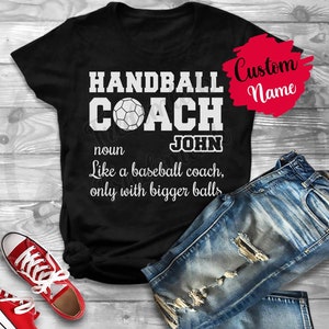 Puede incluir: Camiseta negra con texto blanco que dice "HANDBALL COACH JOHN sustantivo Como un entrenador de béisbol, solo que con pelotas más grandes". La camiseta está sobre una superficie de madera con unos vaqueros azules y unas zapatillas rojas.