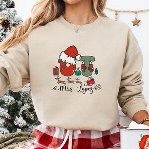 Puede incluir: Sudadera beige con un diseño navideño. El diseño incluye una figura de Papá Noel, la letra "T", mitones, un trineo y renos. El nombre "Mrs. Lopez" está escrito debajo en cursiva.
