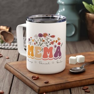 Könnte beinhalten: Weiße Keramik-Reisebecher mit einem bunten Blumenmuster und dem Wort "MEMA" in einer Retro-Schrift. Der Becher hat drei Namen unter dem Wort "MEMA" geschrieben.