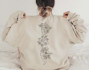 Pode incluir: Uma camisa de moletom bege com um desenho de linha preta de uma espinha dorsal com flores.