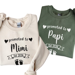 Puede incluir: Dos sudaderas de cuello redondo, una crema y otra verde oliva. La sudadera crema dice "promoted to Mimi Est 2026" con un corazón y un gráfico de huellas. La sudadera verde dice "promoted to Papi Est 2026" con un corazón y un gráfico de huellas.