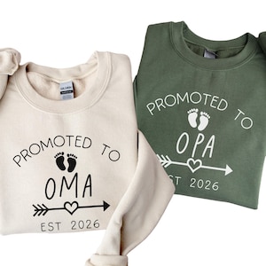 Peut inclure: Deux sweat-shirts col rond, un crème et un vert olive. Le sweat-shirt crème dit "Promu à Oma" avec des empreintes de pas de bébé, une flèche, un cœur et "Est 2026". Le sweat-shirt vert dit "Promu à Opa" avec des graphiques similaires.