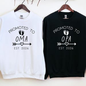 Peut inclure: Deux sweat-shirts à manches longues, un blanc et un noir, sont suspendus côte à côte. Le sweat-shirt blanc dit "Promu à Oma" avec des empreintes de pas de bébé, une flèche, un cœur et "Est 2026". Le sweat-shirt noir dit "Promu à Opa" avec le même design.