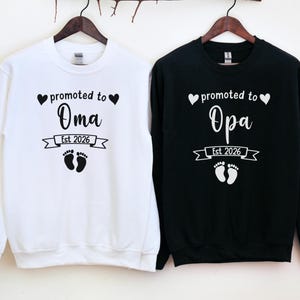 Peut inclure: Deux sweat-shirts à manches longues, un blanc et un noir, suspendus à des cintres en bois. Le sweat-shirt blanc dit "promu à Oma Est 2026" et le sweat-shirt noir dit "promu à Opa Est 2026". Les deux ont un cœur, une bannière et des empreintes de pas de bébé.