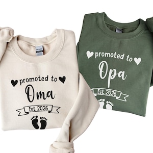 Peut inclure: Deux sweatshirts, un crème et un vert olive, avec les mots "promu à Oma" et "promu à Opa" en écriture noire. Chaque sweat-shirt porte une bannière avec l'inscription "Est 2026" et des empreintes de pas de bébé.