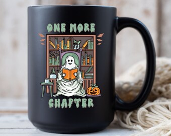 May include: Black ceramic mug with a white interior. The mug features a whimsical illustration of a ghost reading a book in front of a bookshelf, with the text "ONE MORE CHAPTER". A pumpkin is also visible.