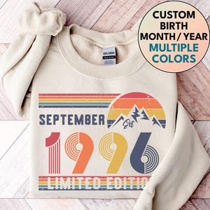 May include: A cream-colored crewneck sweatshirt featuring a graphic print with the text SEPTEMBER 1996 LIMITED EDITION. The design includes a retro-style sunset, mountain silhouettes, and horizontal stripes in shades of red, orange, yellow, and blue.