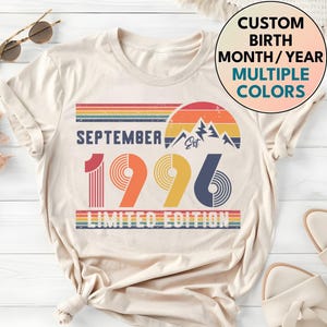 May include: A cream-colored t-shirt featuring a graphic with the text SEPTEMBER 1996 LIMITED EDITION. The design includes a sunset, mountain silhouette, and horizontal stripes in red, orange, yellow, and blue. A circular badge reads CUSTOM BIRTH MONTH/YEAR MULTIPLE COLORS.