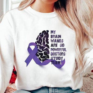 May include: White sweatshirt with a purple ribbon and a black and white brain graphic. The text on the sweatshirt reads "My brain waves are so powerful doctors study them."