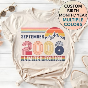 May include: A cream-colored t-shirt featuring a graphic with the text SEPTEMBER 2008 LIMITED EDITION. The design includes a retro-style sunset, mountain silhouettes, and horizontal stripes in shades of orange, yellow, blue, and red.