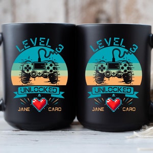 May include: Two black coffee mugs with a retro video game controller design. The mugs say "Level 3 Unlocked" with the names "Jane" and "Caro" on each mug.