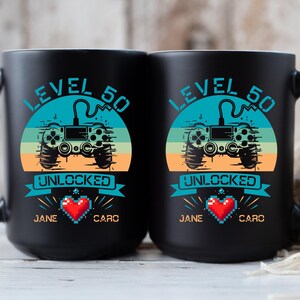 May include: Two black coffee mugs with a retro video game controller graphic and the text "Level 50 Unlocked". The mugs have the names "Jane" and "Caro" printed on them.