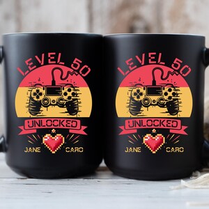 May include: Two black coffee mugs with a retro video game controller design. The mugs have the text "Level 50 Unlocked" and the names "Jane" and "Caro" on them.