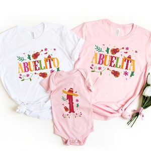 May include: Three pink and white t-shirts with a colourful floral design and the words "Abuelito" and "Abuelita" printed on them. The shirts are for a family of three.