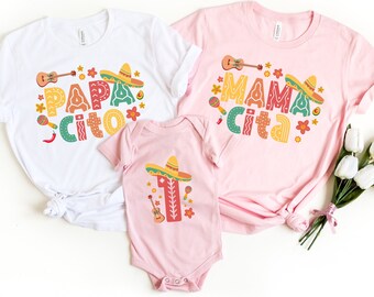 May include: Three matching shirts with a Mexican fiesta theme. The white shirt says "Papa Cito" in colorful letters with a guitar, maracas, and a sombrero. The pink shirt says "Mama Cita" in colorful letters with a guitar, maracas, and a sombrero. The pink baby onesie says "1" in colorful letters with a guitar, maracas, and a sombrero.