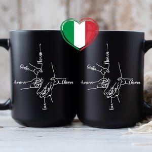 May include: Two black ceramic mugs with white line art depicting hands holding each other. The names "Giulia", "Aurora", "Chiara", and "Luca" are written on the mugs. The word "Nonna" is also written on the mugs. An Italian flag heart is above the mugs.