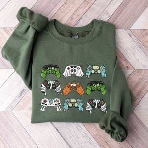 May include: An olive green crewneck sweatshirt featuring a graphic print of nine video game controllers designed with various themes, including a mummy, a ghost, a pumpkin, a skull, and monster-inspired patterns in multiple colors.