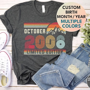 May include: A dark grey heather t-shirt featuring a retro-style graphic with the text OCTOBER 2008 LIMITED EDITION, a mountain silhouette, and horizontal stripes in orange, yellow, and blue. A circular overlay reads CUSTOM BIRTH MONTH/YEAR MULTIPLE COLORS.