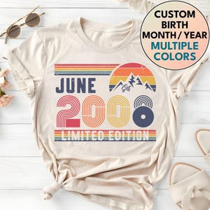 May include: A cream-coloured t-shirt with a retro design. The design includes the text "JUNE 2000 LIMITED EDITION", a rainbow stripe, and a mountain graphic. The text "CUSTOM BIRTH MONTH/YEAR MULTIPLE COLORS" is also visible.