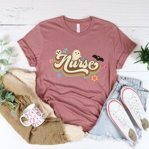 Op de afbeelding: Een mauvekleurig T-shirt met het woord "Nurse" in een retro lettertype, vergezeld van een spook, vleermuis en bloemenafbeeldingen. Het shirt is gestyled met een spijkerbroek en witte sneakers, wat een casual look suggereert.