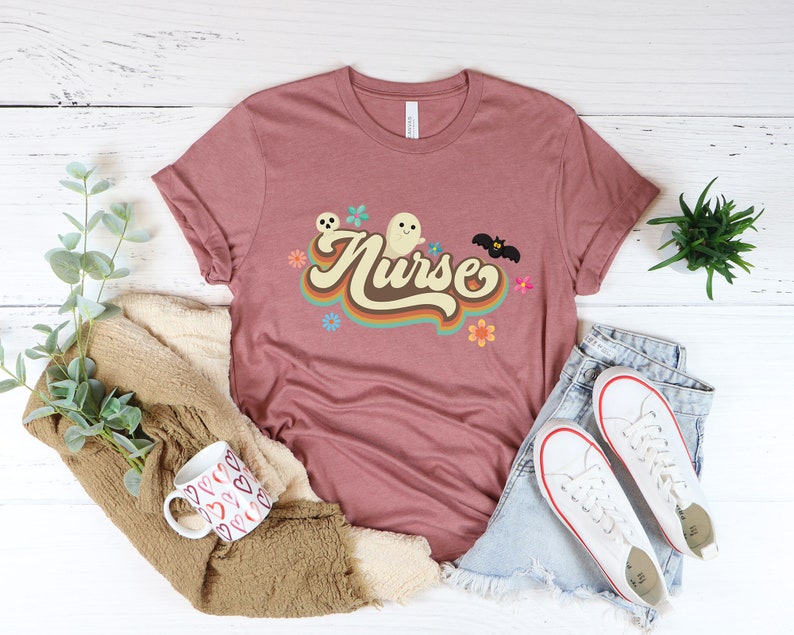 Op de afbeelding: Een mauvekleurig T-shirt met het woord "Nurse" in een retro lettertype, vergezeld van een spook, vleermuis en bloemenafbeeldingen. Het shirt is gestyled met een spijkerbroek en witte sneakers, wat een casual look suggereert.