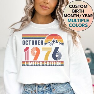 May include: A white crewneck sweatshirt featuring a graphic print with the text OCTOBER 1976 LIMITED EDITION. The design includes a retro-style sunset, mountain silhouettes, and striped horizontal lines in shades of red, orange, yellow, and blue.