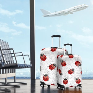 May include: Two white suitcases with ladybug print luggage covers. The larger suitcase has a black handle and wheels. The smaller suitcase has a black handle and wheels. The suitcases are in an airport terminal with a plane in the background.