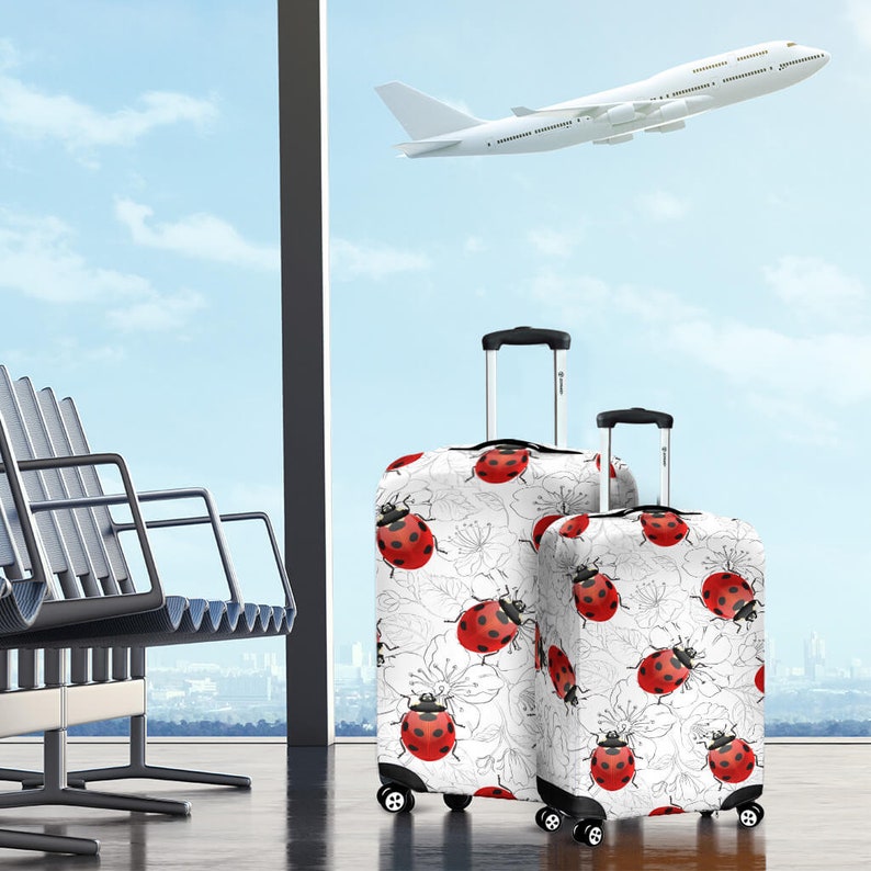 May include: Two white suitcases with ladybug print luggage covers. The larger suitcase has a black handle and wheels. The smaller suitcase has a black handle and wheels. The suitcases are in an airport terminal with a plane in the background.