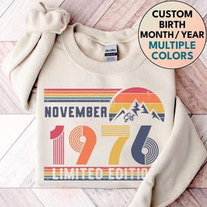 May include: A cream-colored crewneck sweatshirt featuring a graphic print with the text NOVEMBER 1976 LIMITED EDITION. The design includes a retro-style sunset, mountain silhouettes, and horizontal stripes in shades of orange, yellow, and blue.