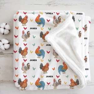 May include: White baby blanket with a repeating pattern of colorful rooster illustrations and the name "JAMES" in black. The blanket is folded, revealing a soft, white interior. Decorative cotton buds are in the background.