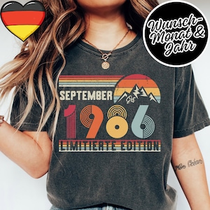 May include: A charcoal gray t-shirt featuring a retro-style graphic with a sunset, mountain silhouette, and the text SEPTEMBER 1986 EST LIMITIERTE EDITION. The design uses horizontal stripes and circular patterns in shades of orange, yellow, and blue.
