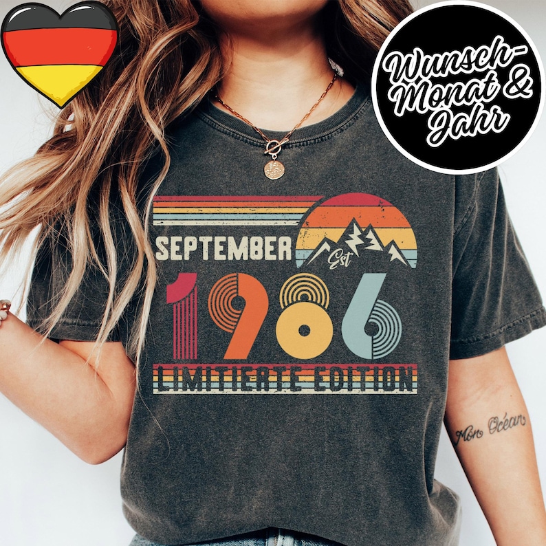May include: A charcoal gray t-shirt featuring a retro-style graphic with a sunset, mountain silhouette, and the text SEPTEMBER 1986 EST LIMITIERTE EDITION. The design uses horizontal stripes and circular patterns in shades of orange, yellow, and blue.